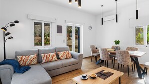 Ein modernes Wohnzimmer mit grauem Sofa, gemusterten Kissen, einem Holztisch und einem Essbereich mit Tisch und Stühlen.