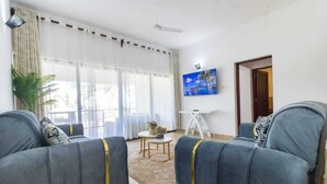 A living room with two grey armchairs, a small round table with a plant, and a painting on the wall. - Lux Suites Vee Xanadu Luxury Apartments (Mombasa)