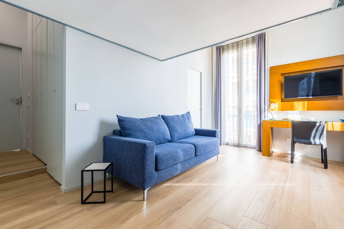 Un salon moderne avec un canapé bleu, une petite table d’appoint noire, un bureau en bois avec une chaise et un téléviseur fixé au mur.