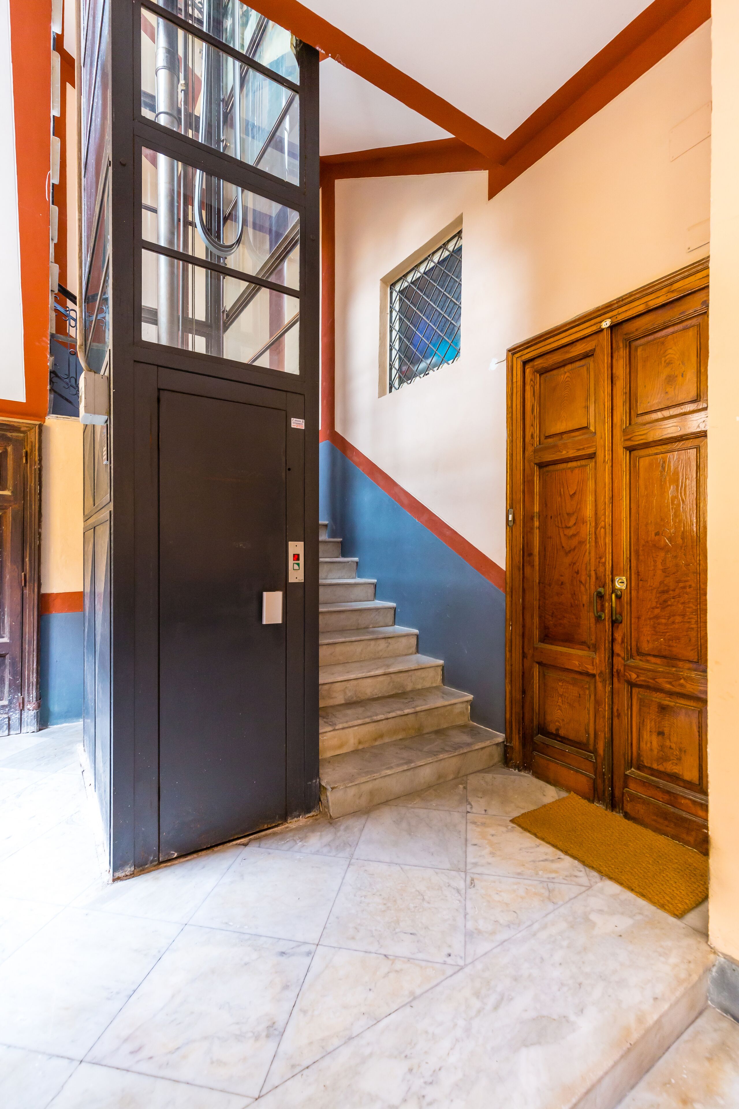 A staircase with a black elevator door, a wooden door, and a small window with a metal grille.