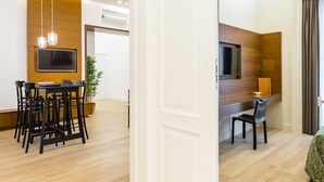 Una habitación de hotel moderna con suelo de madera, una zona de comedor con mesa y sillas, un televisor montado en la pared y un escritorio con una silla.