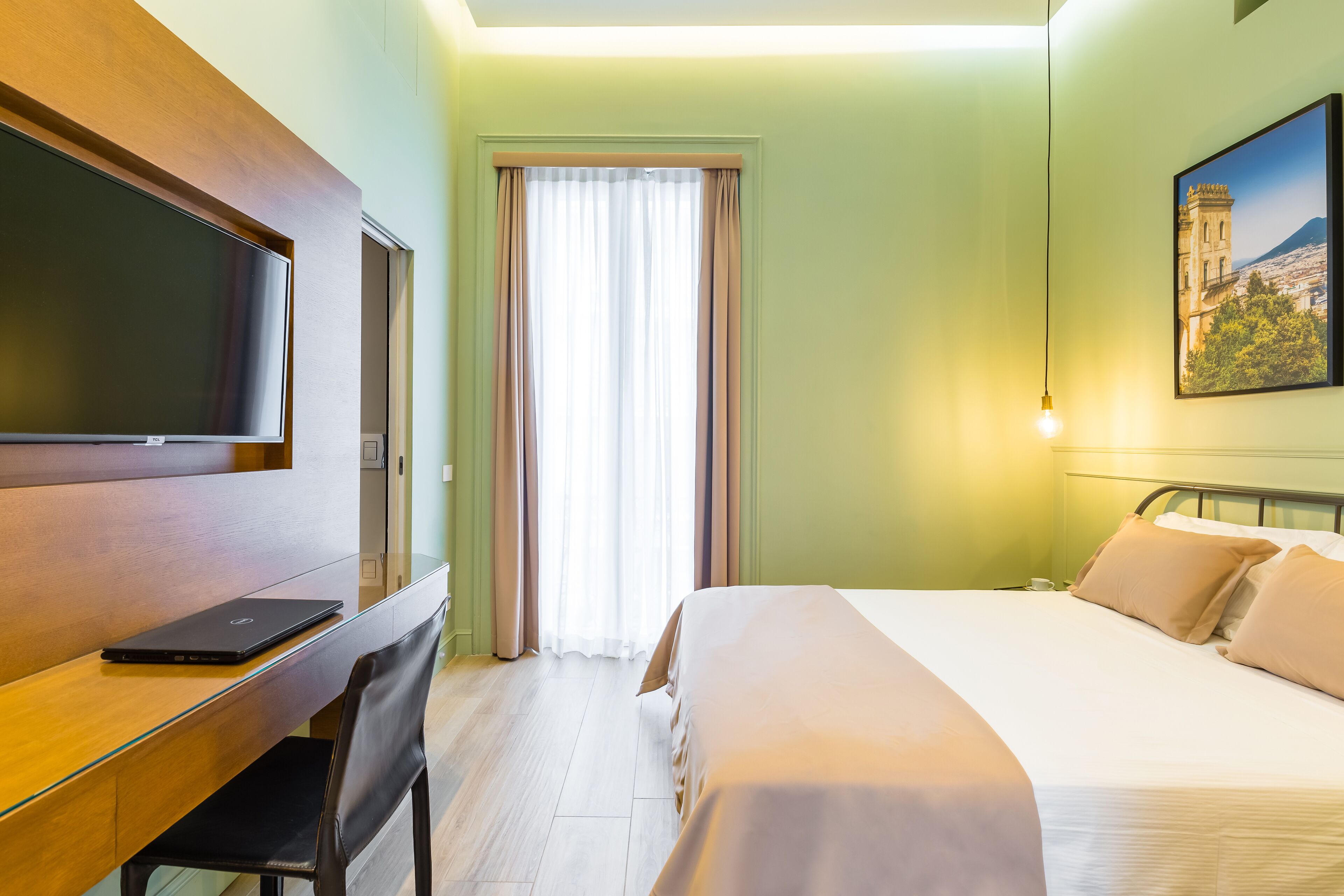 Ein Hotelzimmer mit einem Bett, einem Schreibtisch mit einem Laptop, einem Fernseher und einem Bild an der Wand.