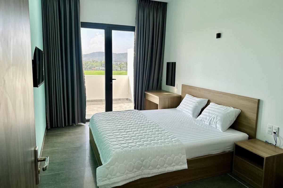 Una habitación de hotel con una cama grande, un cabecero de madera, una mesita de noche y vistas al exterior a través de una puerta corredera.