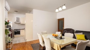 A modern living room with a glass dining table, white chairs, a black leather sofa, and a small kitchen area in the background. - Coastal Comfort Apartment (Podstrana)