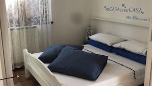 Une chambre à coucher avec un lit blanc, des oreillers bleus, une tête de lit blanche, un luminaire fixé au mur et un panneau décoratif.