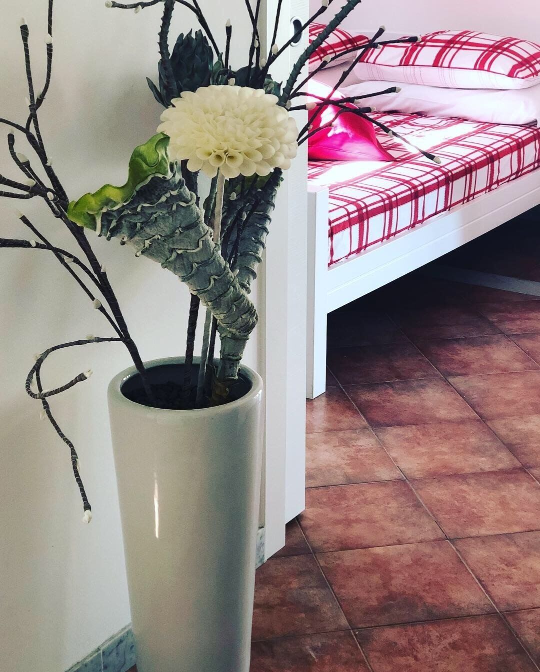 A room with a bed, a white vase with a plant, and a tiled floor.