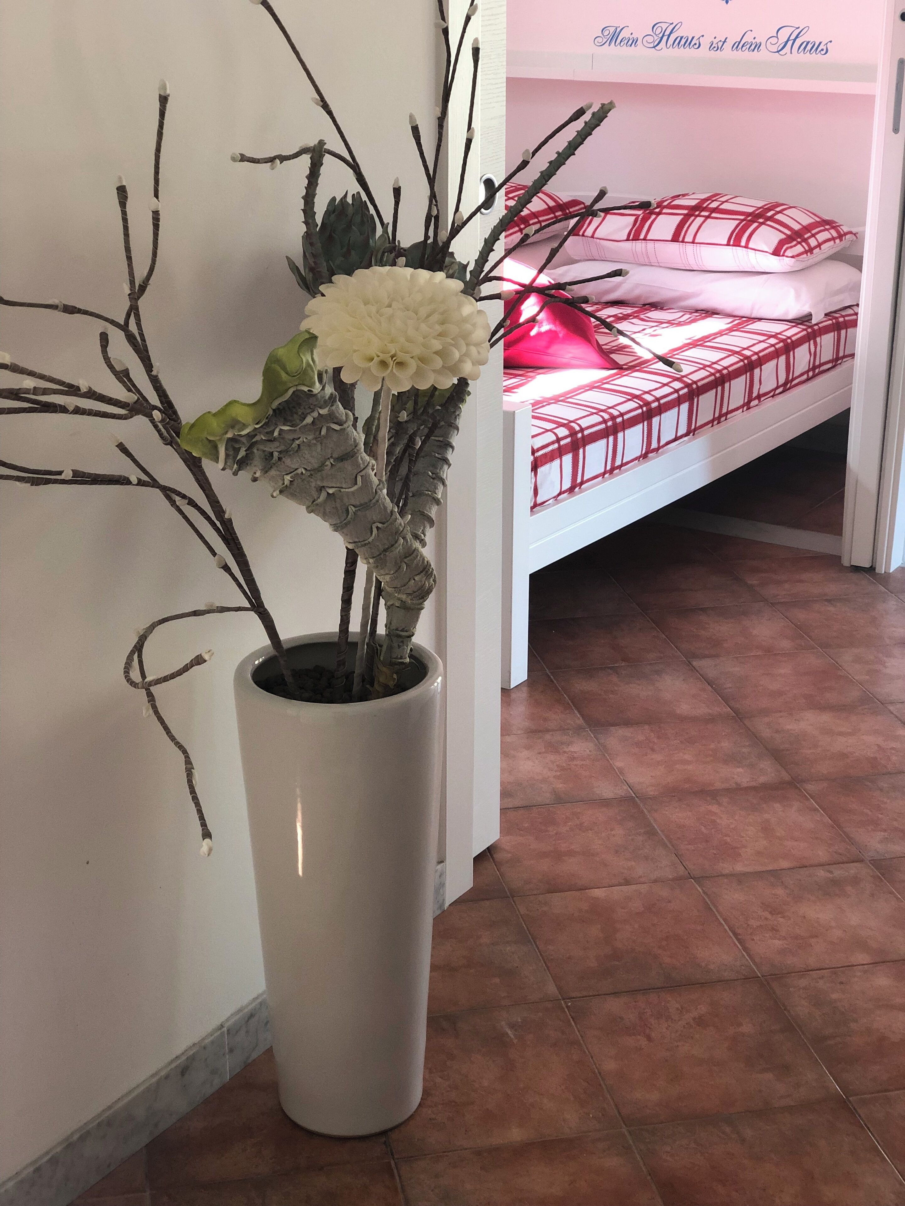 Une plante en pot, aux feuilles et aux fleurs blanches, se trouve dans une pièce où il y a un lit et un mur rose.