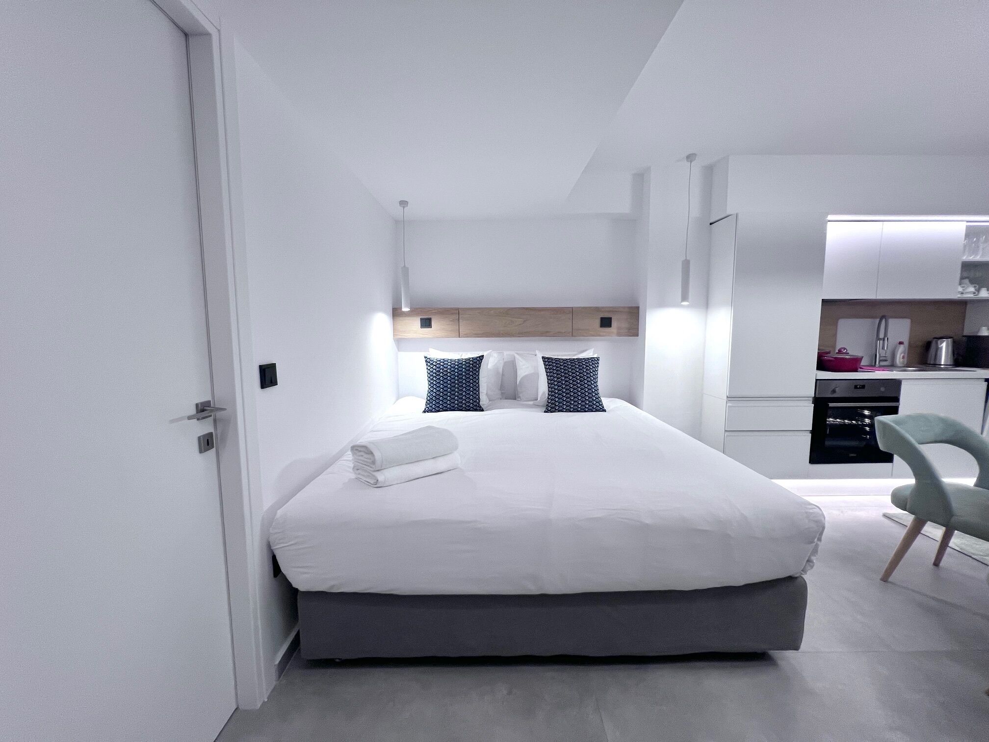 Moderni hotellihuone, jossa on siististi pedattu sänky, pieni keittokomero ja harmaa tuoli.