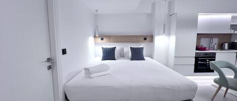 A modern hotel room with a neatly made bed, a small kitchenette, and a grey chair.