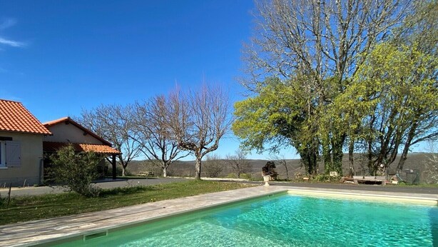 Une piscine aux eaux cristallines bordée d’une terrasse en bois, une maison avec un toit rouge et des arbres en arrière-plan.