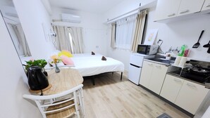 Une pièce compacte et bien éclairée, comprenant un lit, une petite table à manger avec des chaises, un coin cuisine et une fenêtre avec des rideaux.