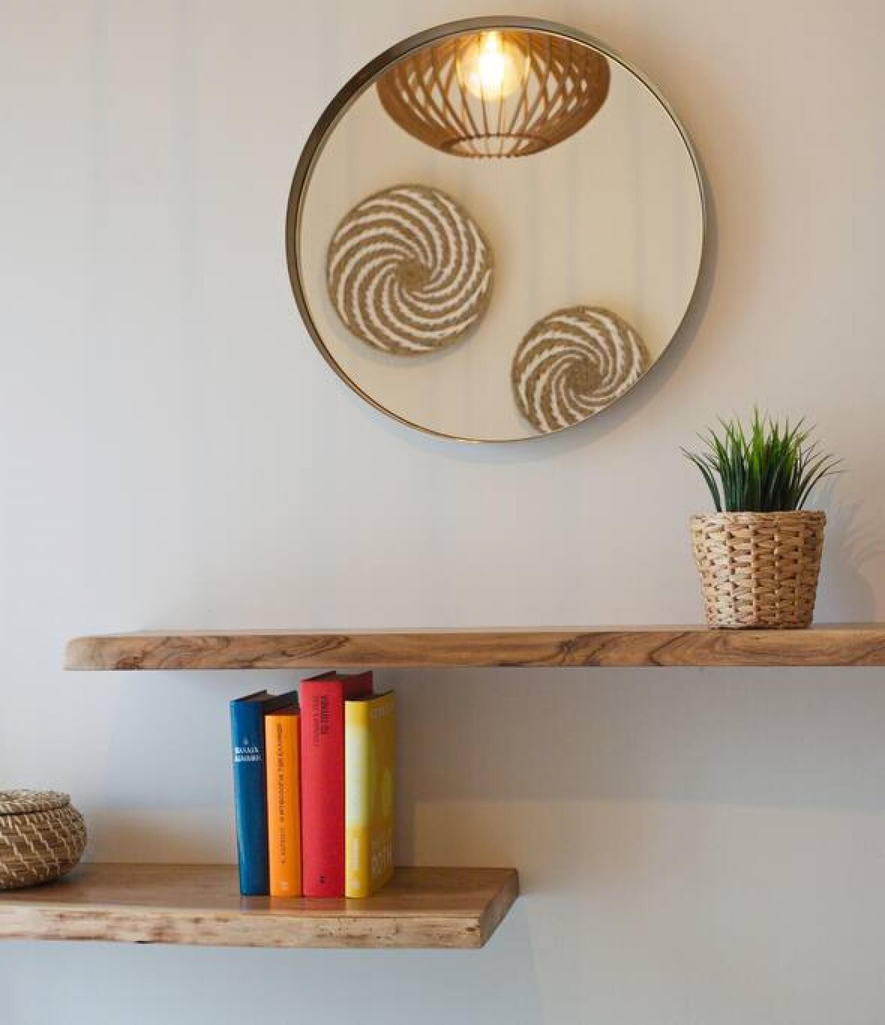 A wall-mounted circular mirror with a light fixture, two wooden shelves with books and a woven basket with a plant.