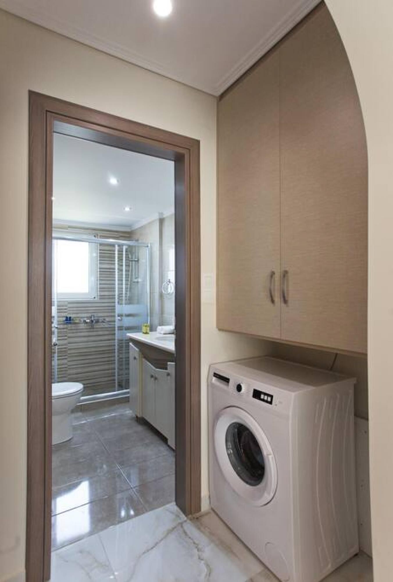A compact bathroom with a glass shower enclosure, a washing machine, and a wooden cabinet.