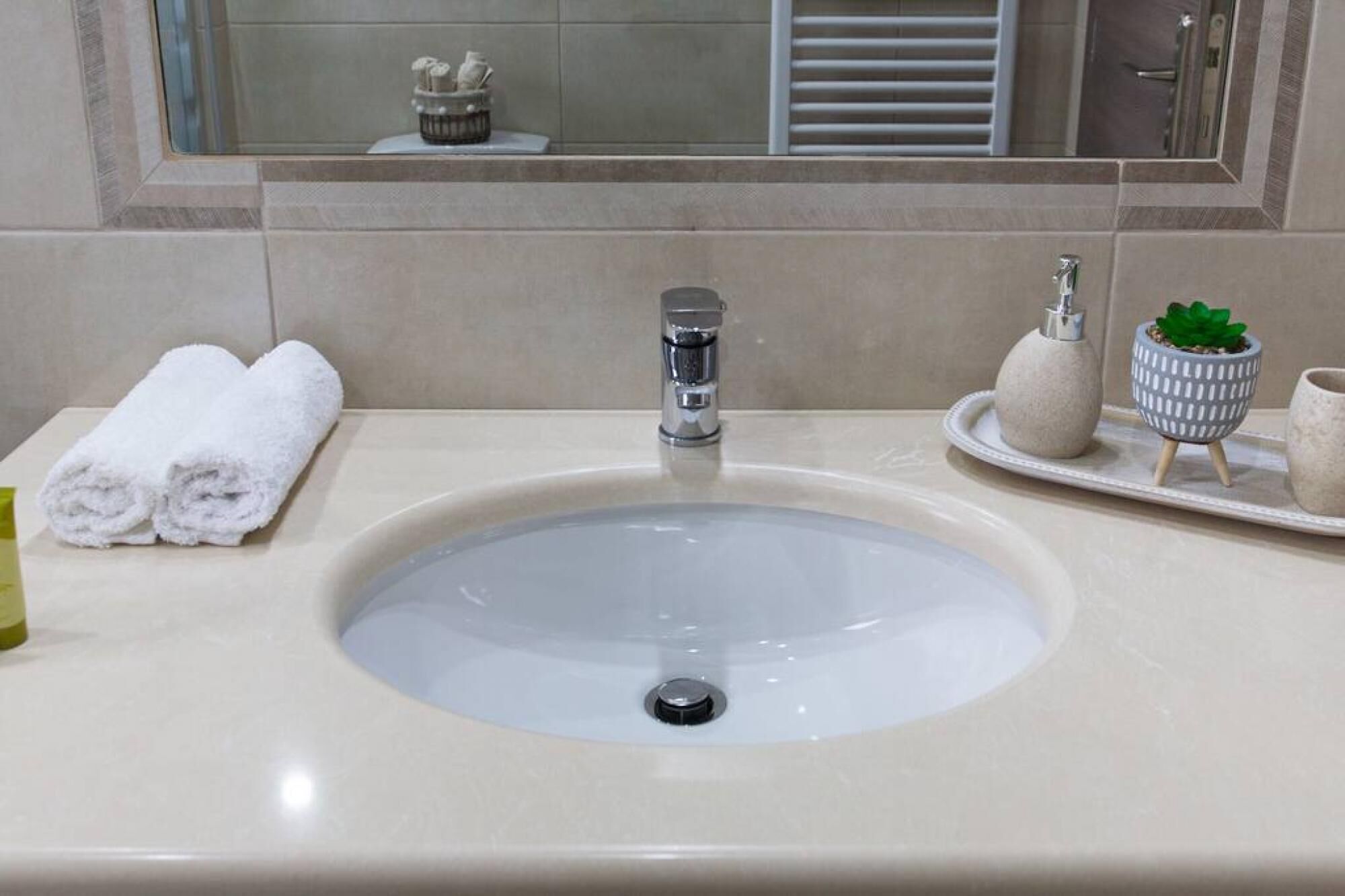 A bathroom sink with a mirror above it, a towel rack with towels, and a tray with a plant and a soap dispenser.