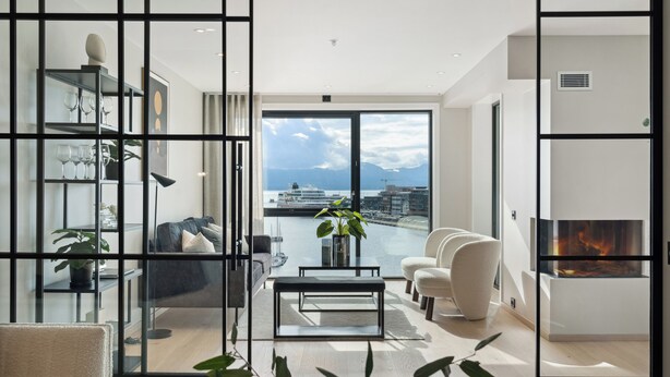 A modern living room with a large window, a fireplace, and a view of a harbor. - TotalApartments Vervet Fram F (Tromsø)