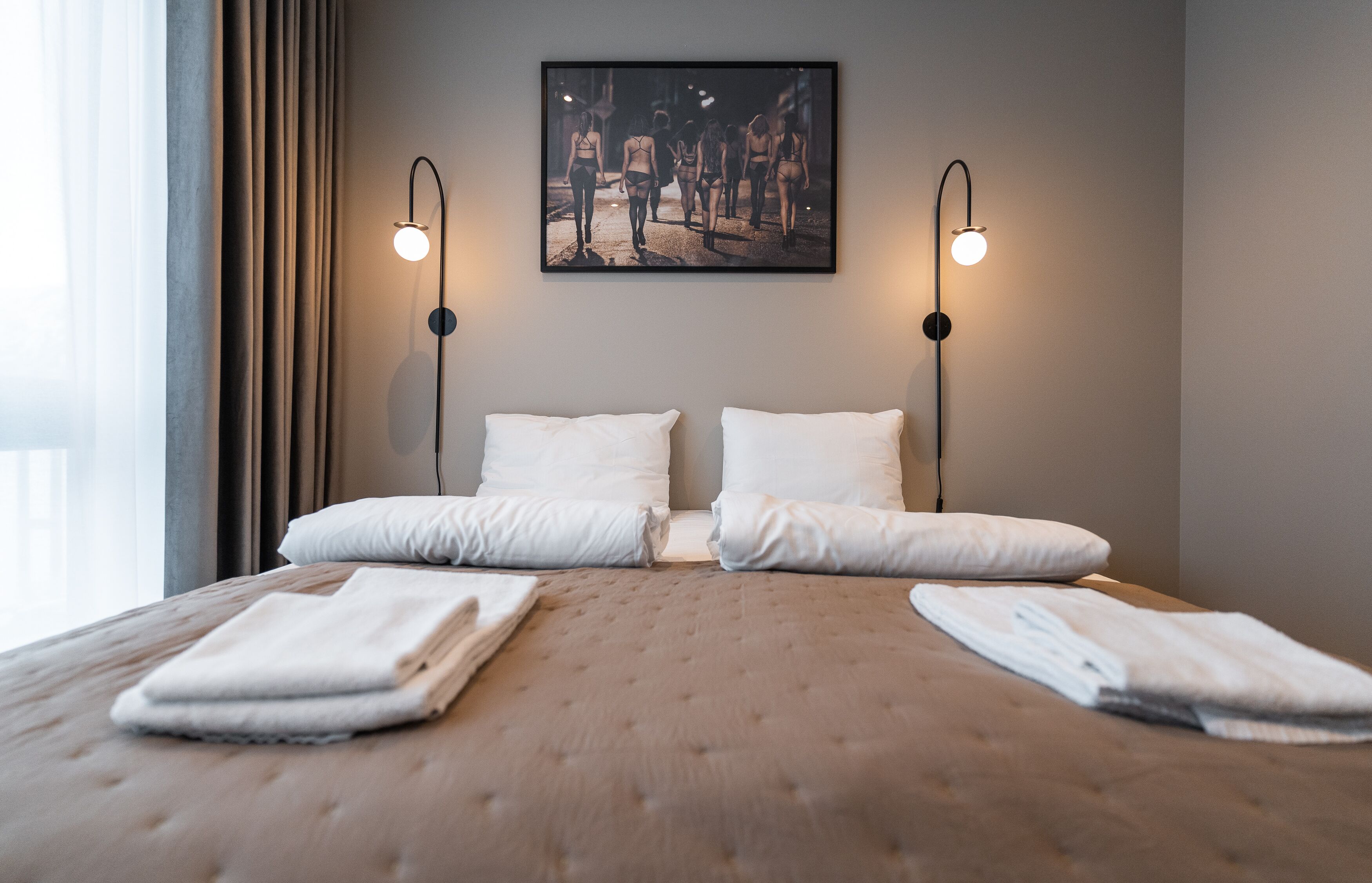 A neatly made bed with two pillows and folded towels, a framed picture on the wall, and two wall-mounted lamps.