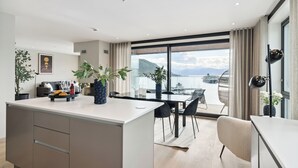 A modern kitchen with a large island, dining area, and large windows offering a view of the sea. - TotalApartments Vervet Fram F (Tromsø)
