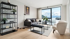 A modern living room with a sofa, a coffee table, and a bookshelf. There is a large window with a view of the sea and boats. - TotalApartments Vervet Fram F (Tromsø)