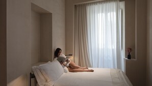 Una habitación con una cama, una ventana con cortinas translúcidas y un pequeño estante con un florero.