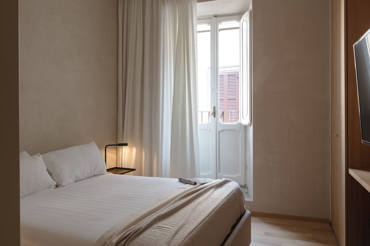 A bedroom with a bed, a nightstand, a lamp, a TV, and a window with curtains.