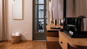 Quarto de hotel com piso de madeira, espelho, cesta com toalhas, cafeteira e janela com persianas.