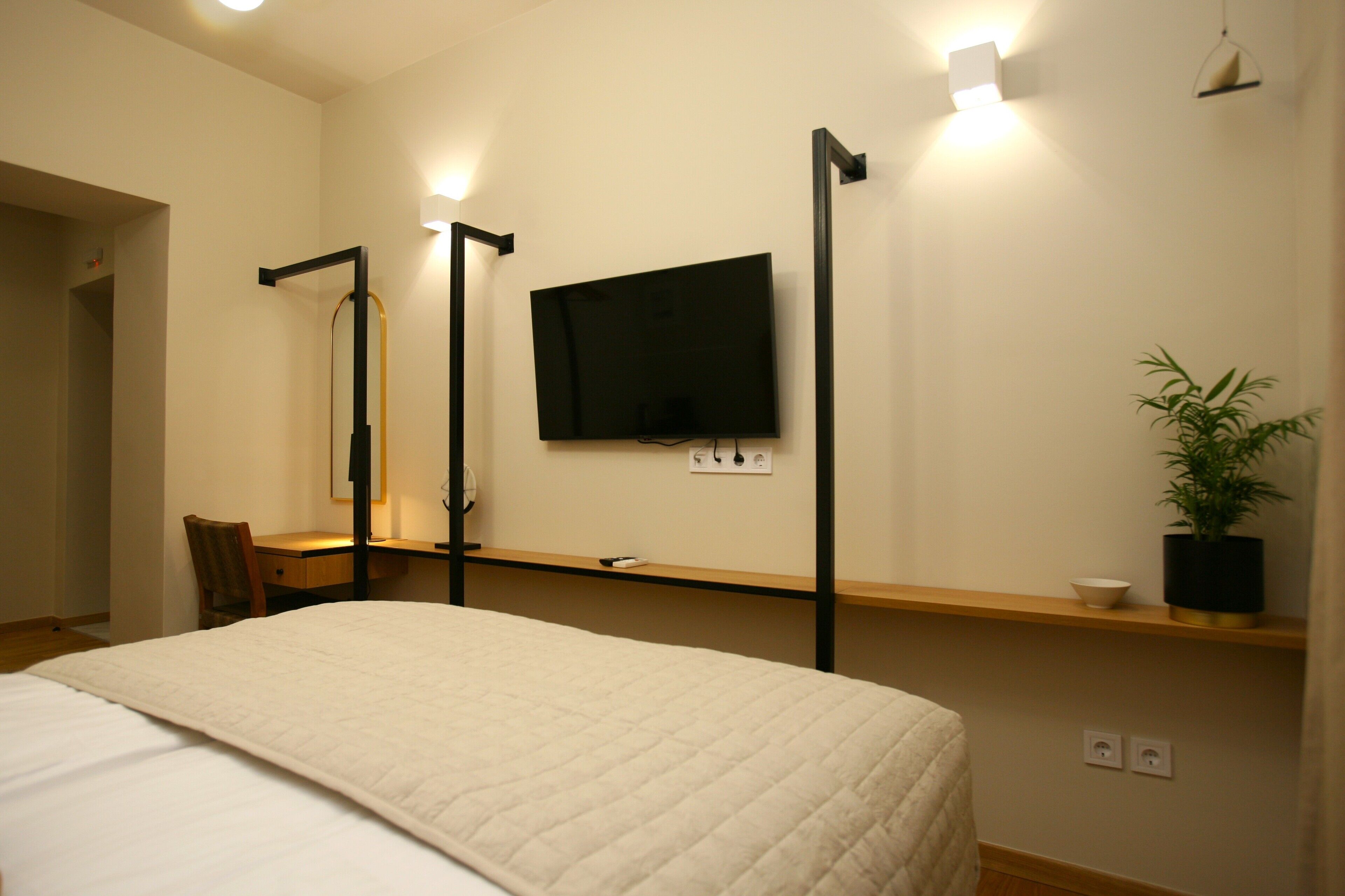 A hotel room with a bed, a desk, a chair, a television mounted on the wall, a potted plant, and a shelf with a bowl.