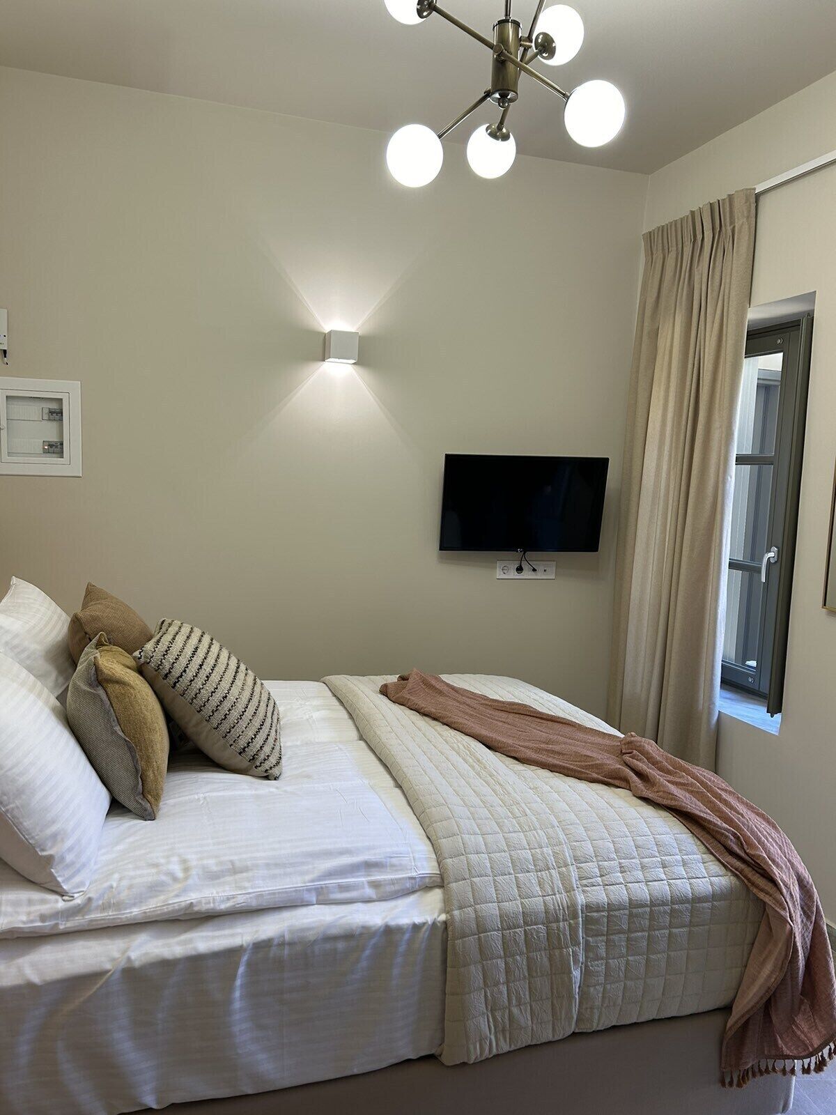 A bedroom with a bed, pillows, a blanket, a TV, and a window with curtains.
