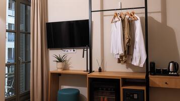 Una habitación de hotel moderna con un ventanal grande, un televisor de pantalla plana, un escritorio de madera con una planta, un pouf color turquesa, un minibar y un perchero.