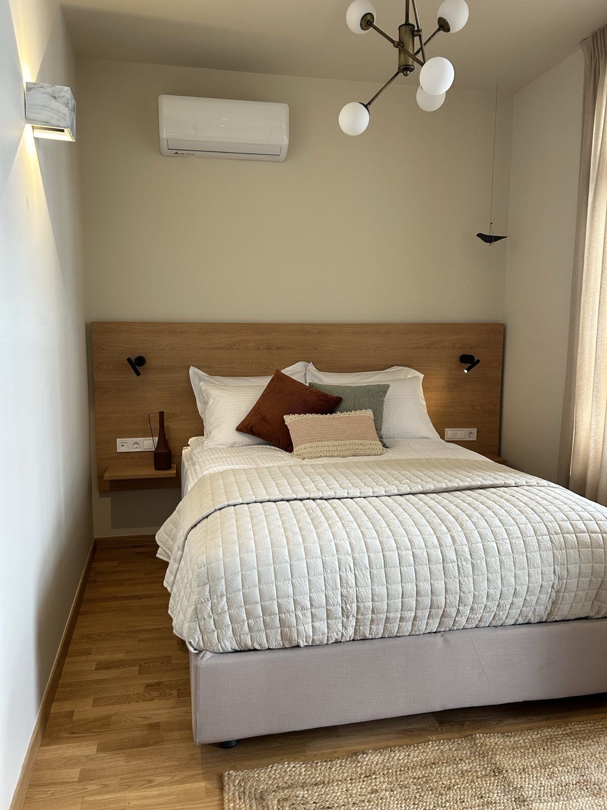 Un lit bien fait, avec une couette blanche matelassée, une tête de lit en bois et une table de chevet surmontée d’une lampe.