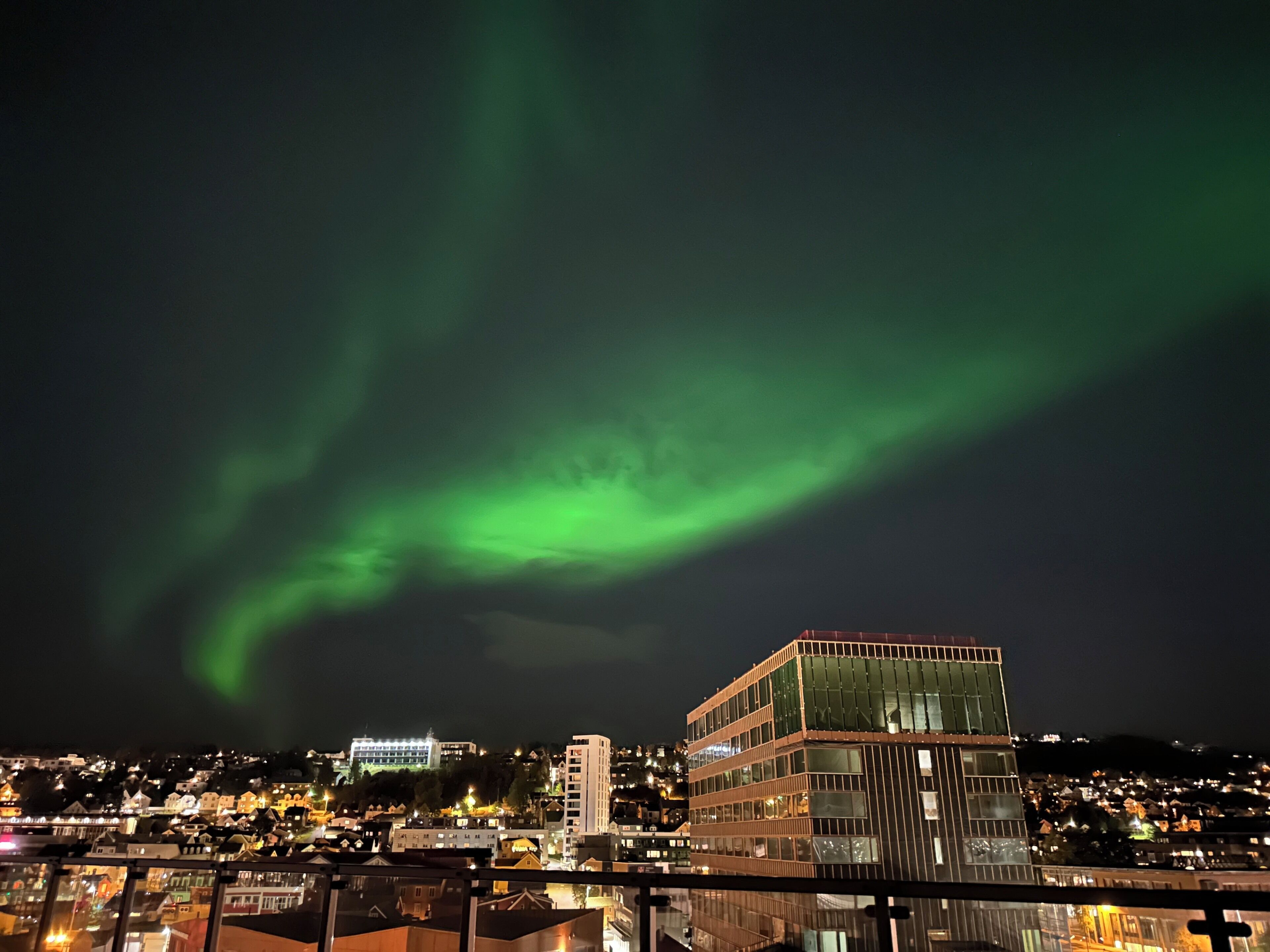 A cityscape at night with a prominent building featuring a glass facade and a green aurora in the sky.