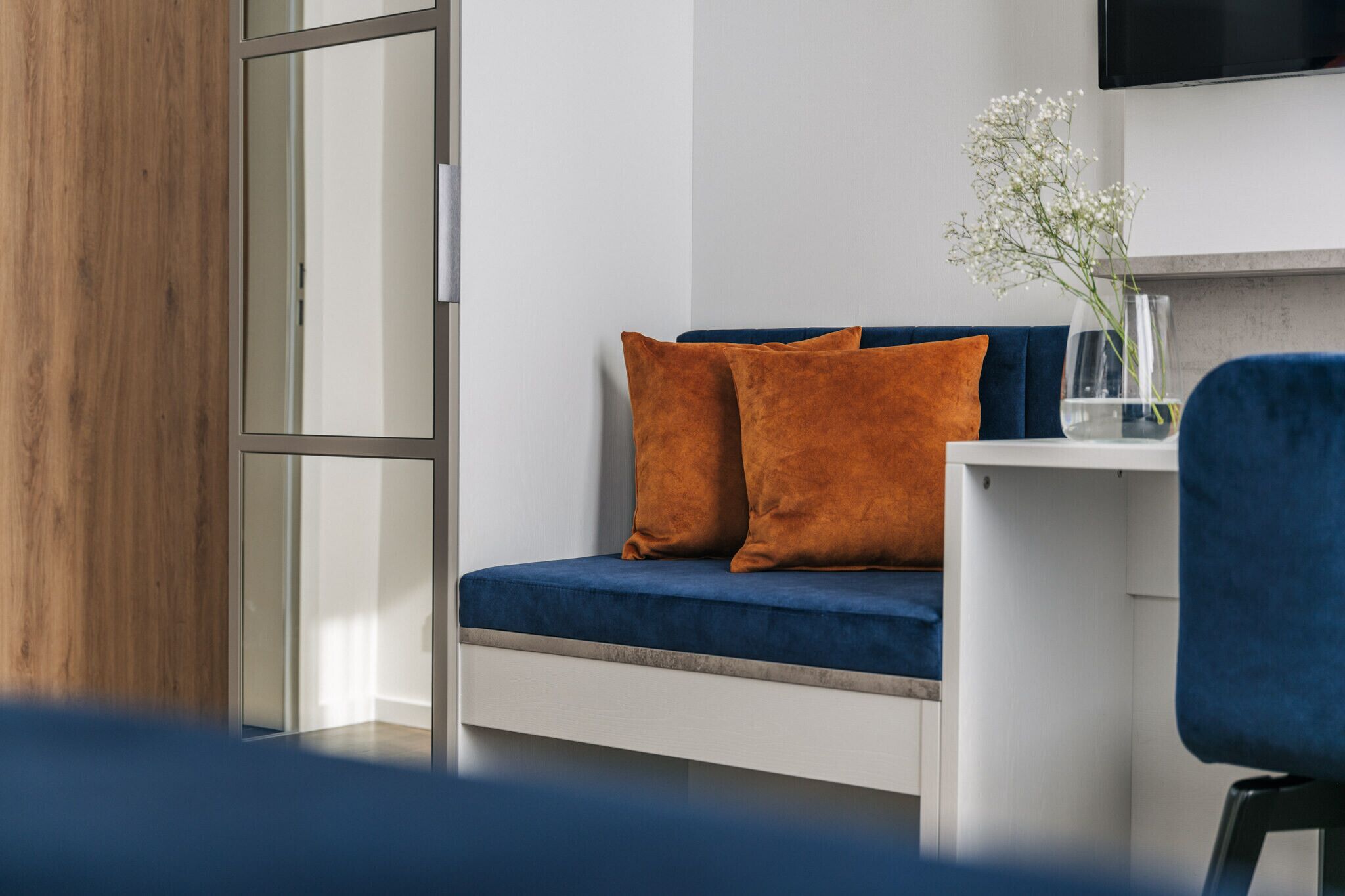 A modern room with a blue sofa, two orange cushions, a small table with a vase of flowers, and a TV mounted on the wall.