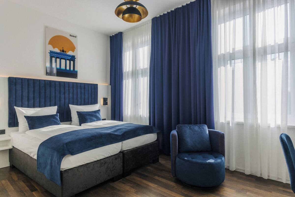 A modern hotel room with a large bed, a blue armchair, and a view of the cityscape through sheer curtains.