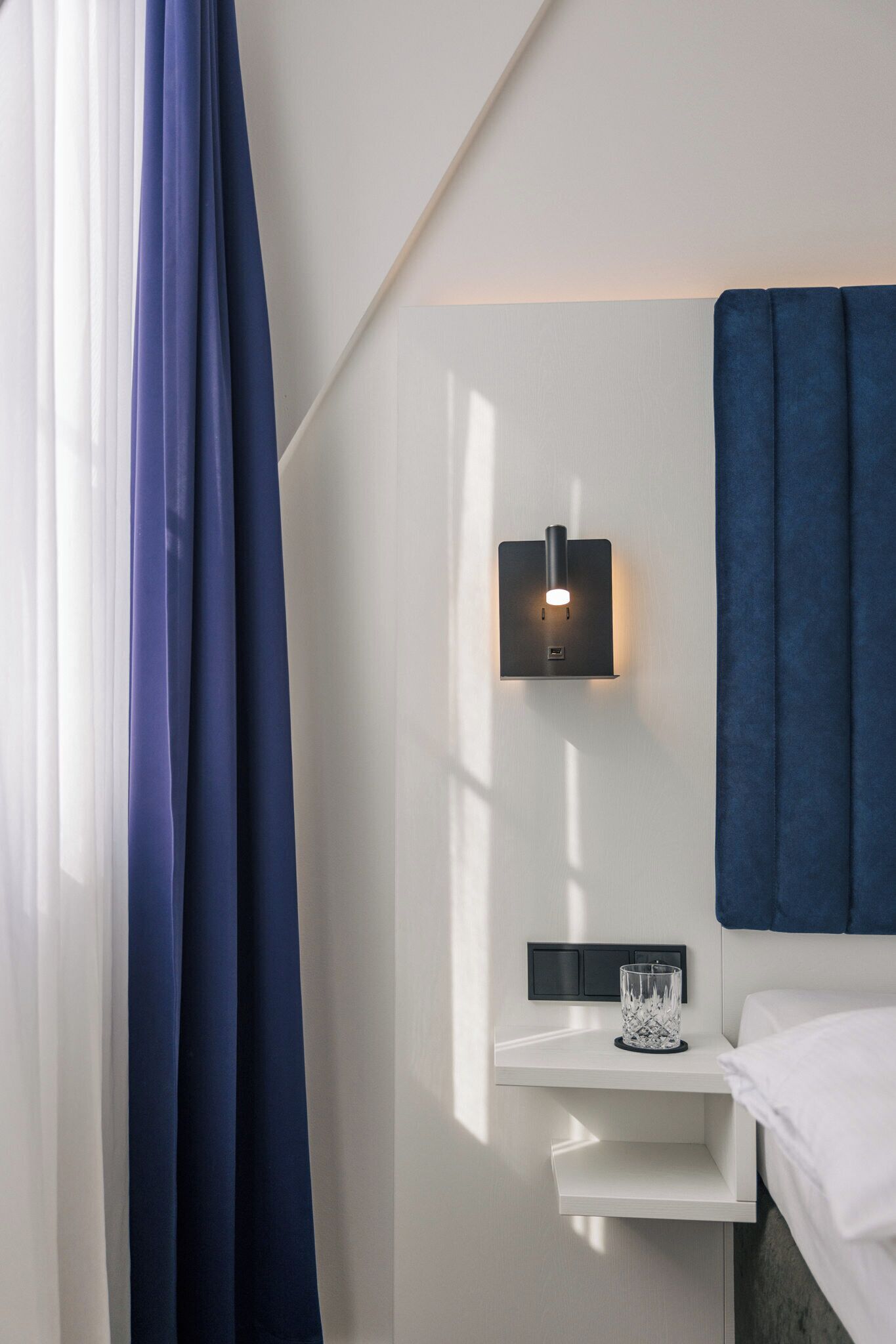 A modern hotel room with a white wall, a blue headboard, and a bedside table with a glass container.