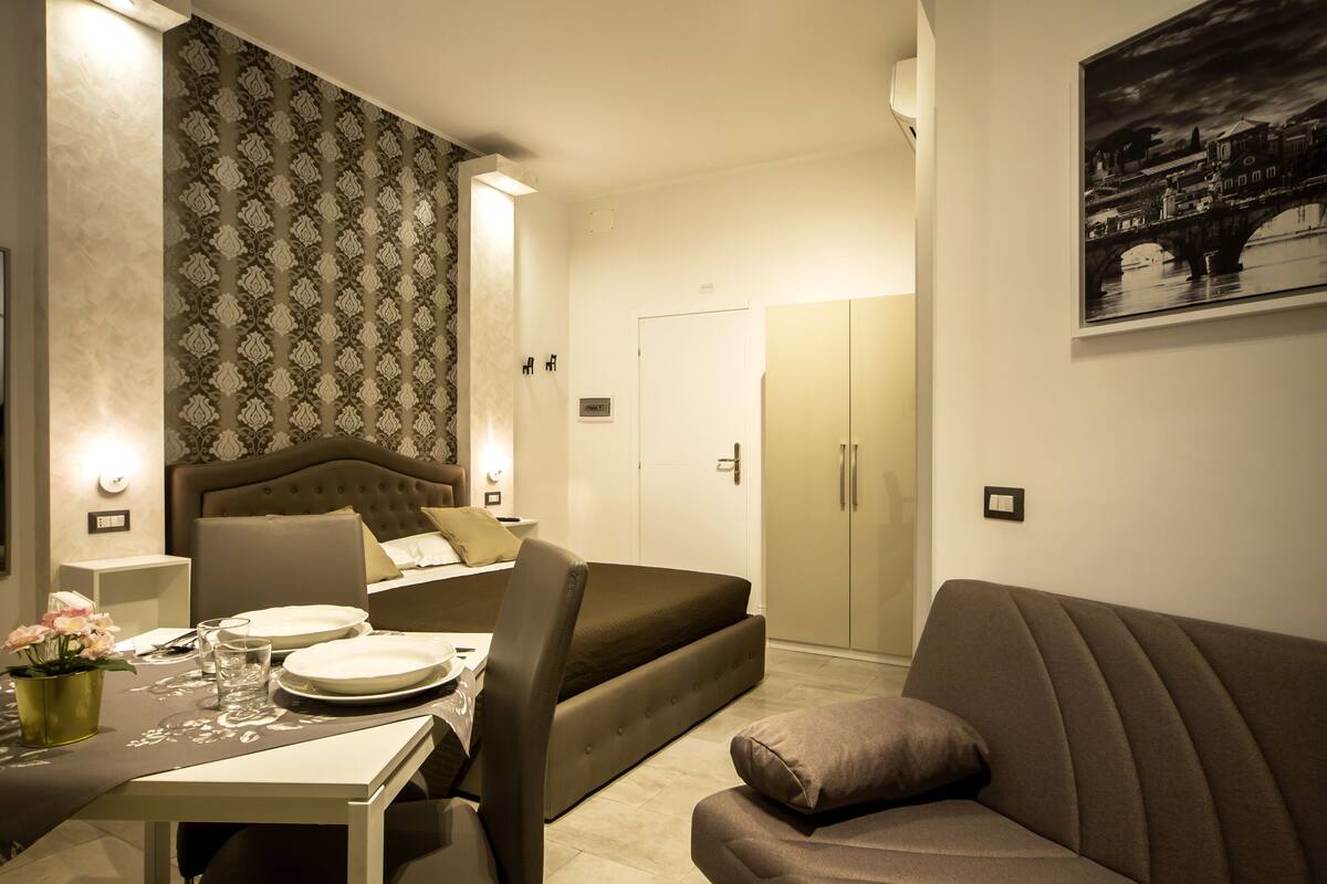 A compact hotel room with a bed, a sofa, a dining table with plates and glasses, and a patterned wallpaper.