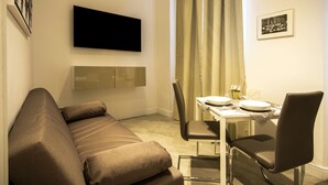 A modern hotel room with a sofa, a dining table set for two, a flat-screen TV mounted on the wall, and a framed picture on the wall. - Residenza Tupini S.Pietro Suites (Rome)