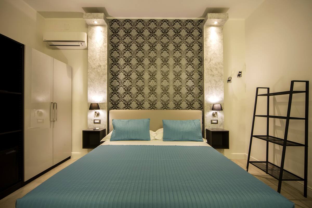 A bedroom with a large bed, two bedside tables with lamps, a wardrobe, and a wall-mounted shelf.