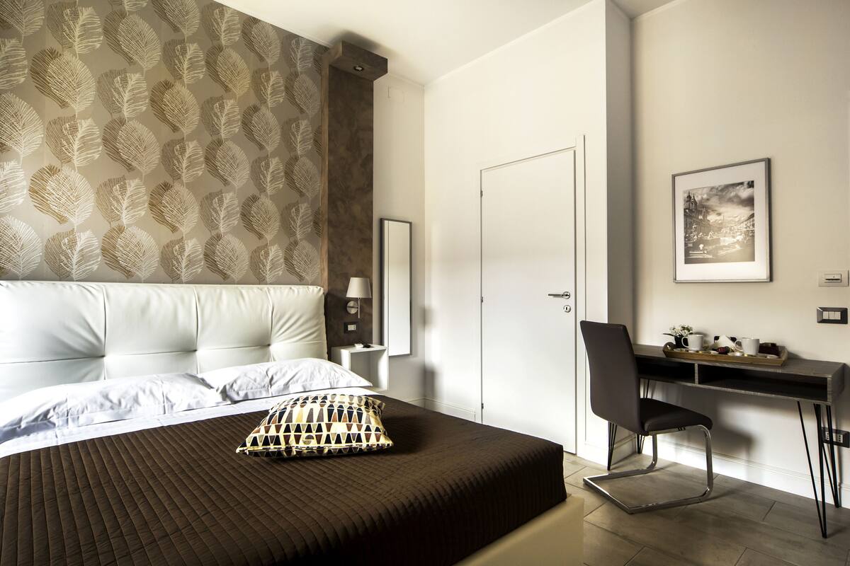 A hotel room with a bed, a desk, and a chair. There is a wall with a leaf pattern and a framed picture on the wall.