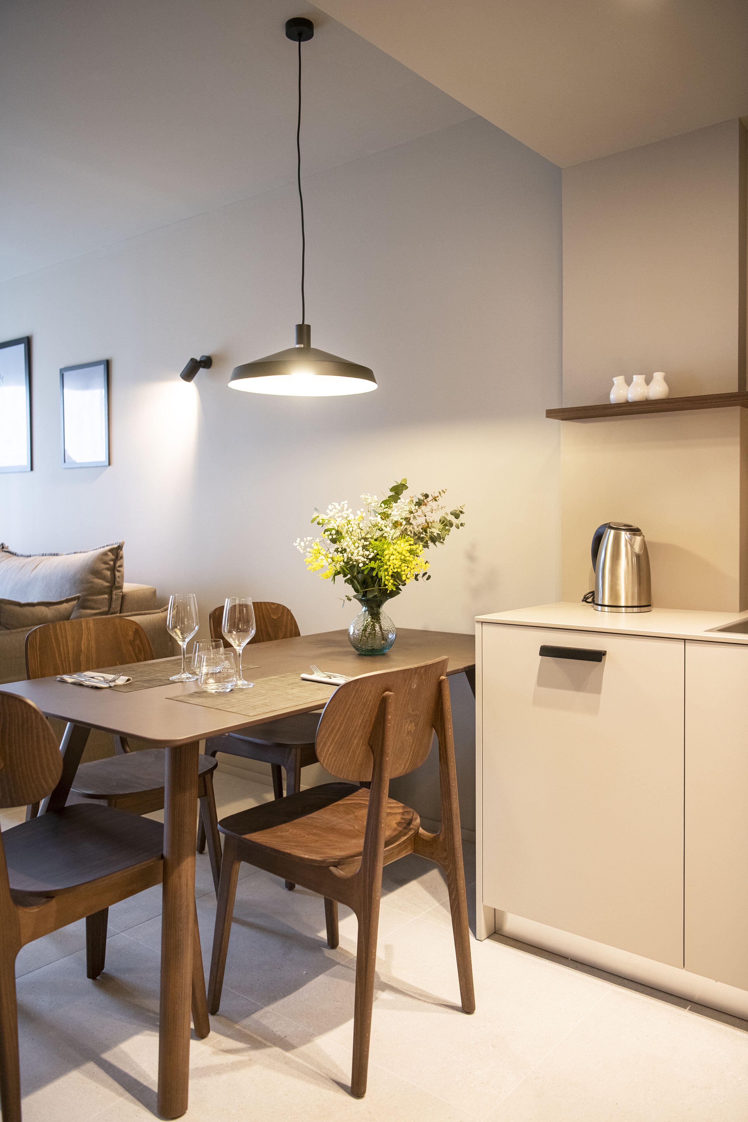 A modern dining area with a wooden table, chairs, a vase with flowers, and a kettle on a cabinet.
