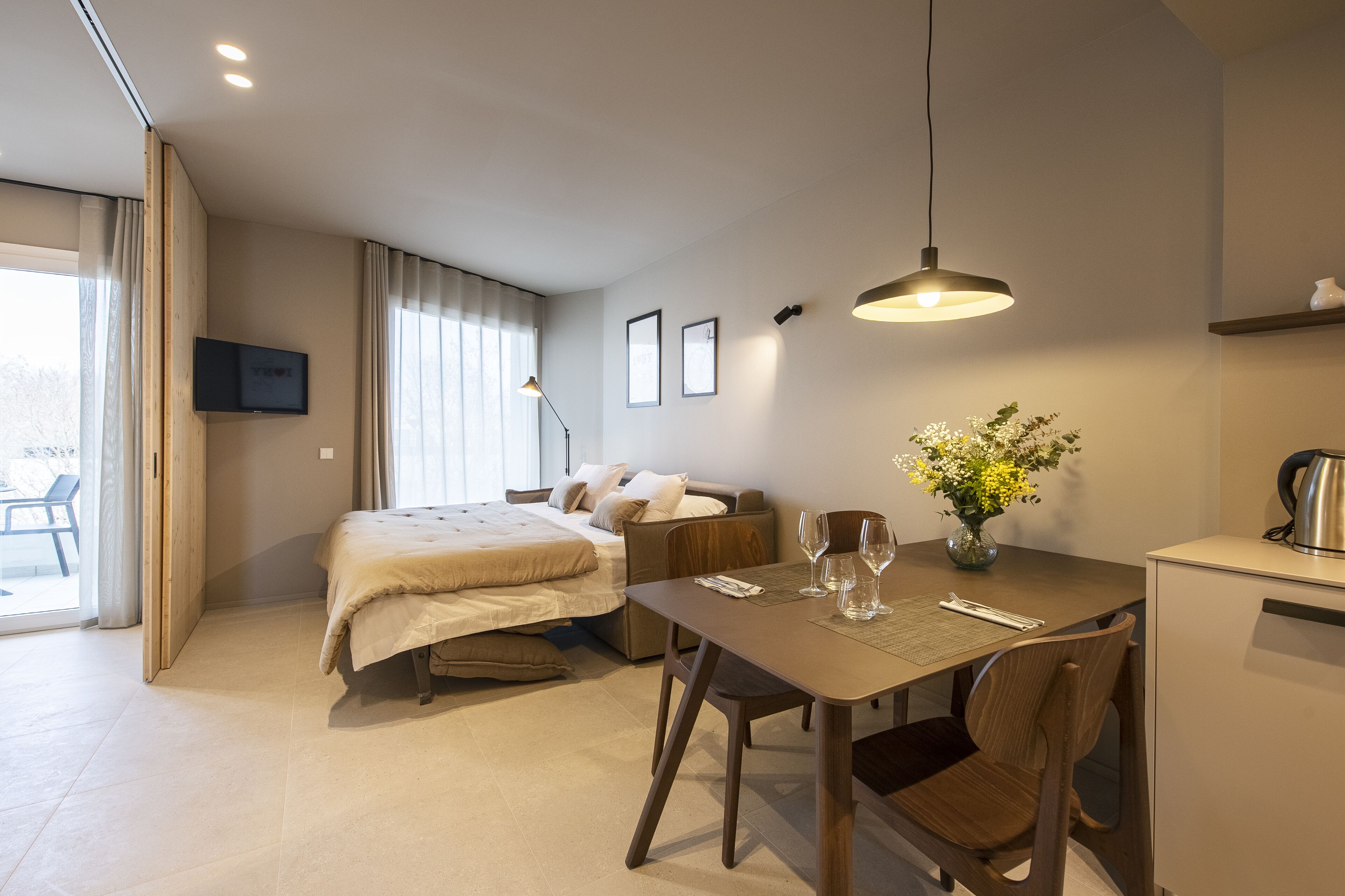 Una habitación de hotel moderna con una cama, una mesa de comedor con sillas, una pequeña cocina y un televisor montado en la pared.