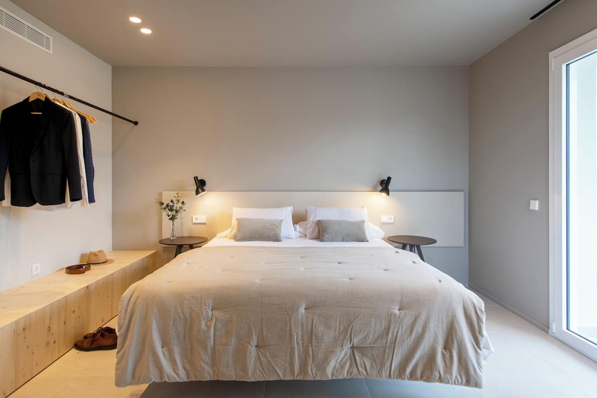 Une chambre moderne avec un grand lit, des tables de chevet, un banc avec des chaussures, un porte-manteau et une grande fenêtre.