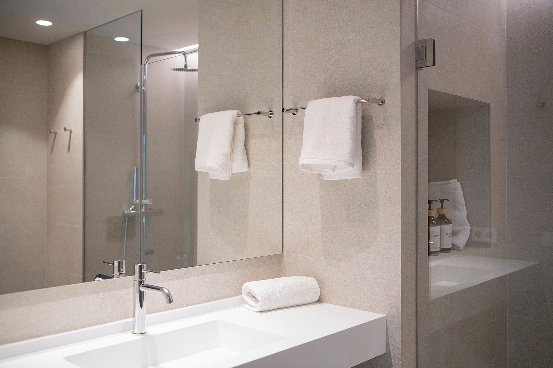 Ein modernes Badezimmer mit einer in Glas abgeschlossenen Dusche, einem weißen Waschbecken und zwei Handtüchern, die auf einem Haken/Gestell hängen.