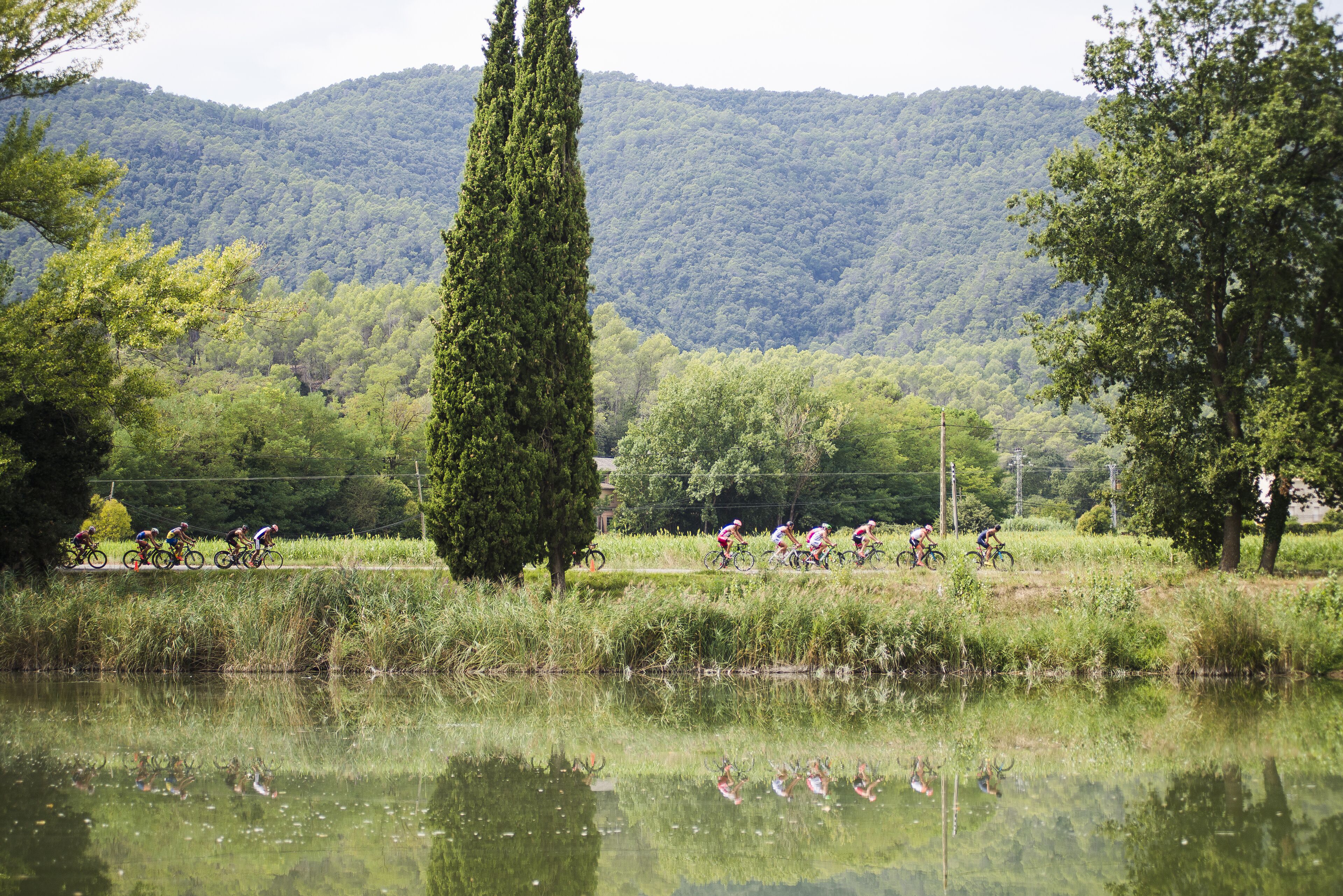 Un grupo de ciclistas en un sendero cerca de una masa de agua, con una colina boscosa al fondo.