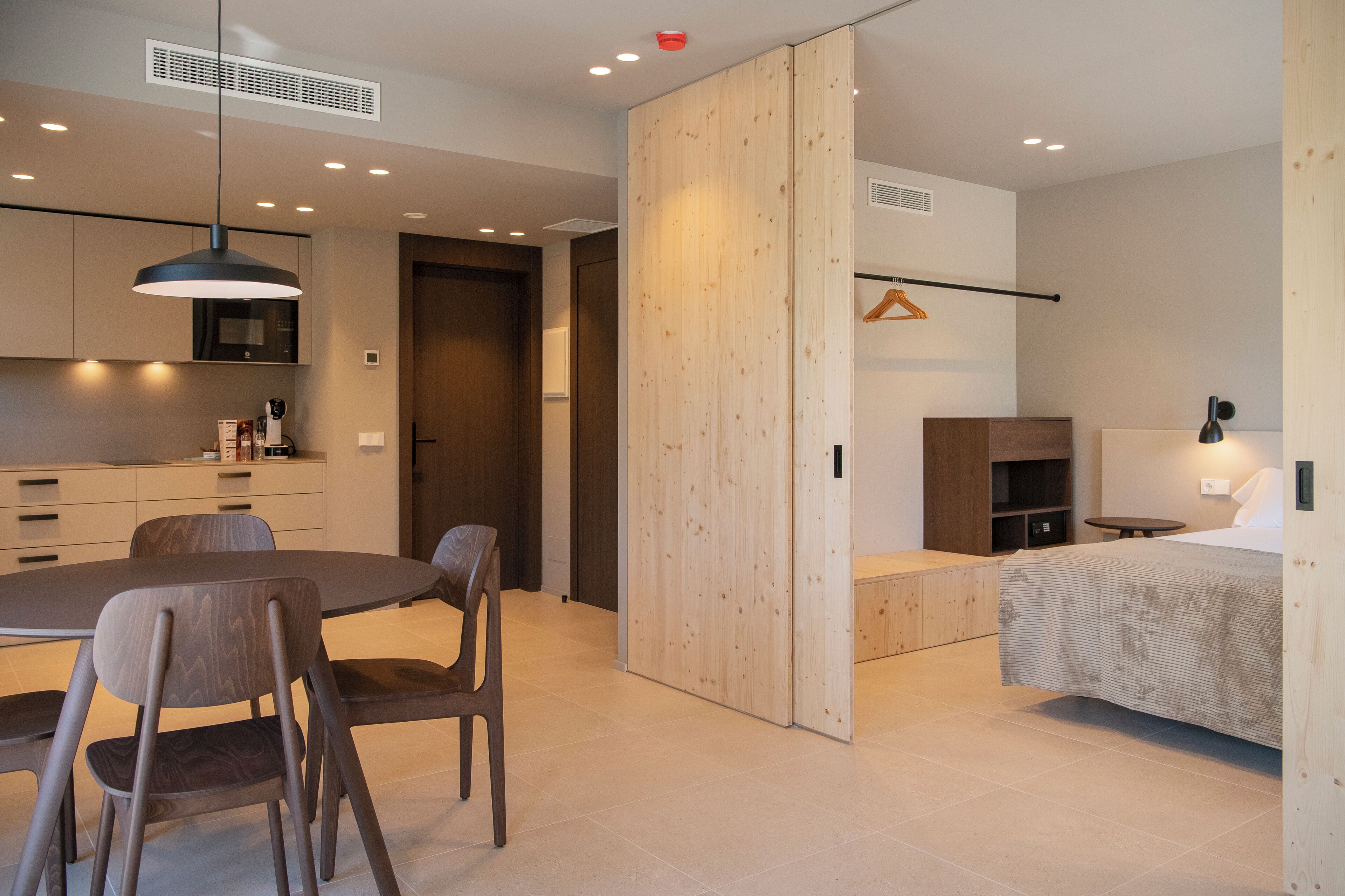 Una habitación moderna y luminosa con zona de comedor, cama y una mesita de noche con lámpara.