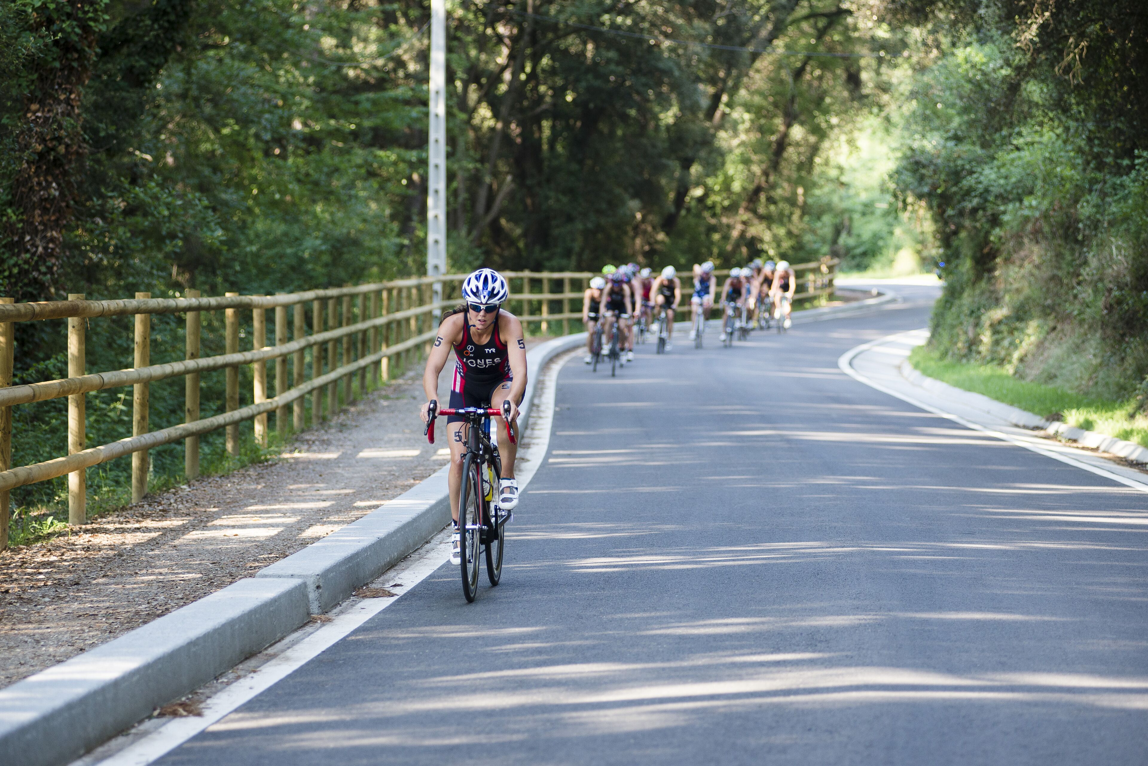 Un gruppo di ciclisti su una strada asfaltata, con una recinzione in legno da un lato e alberi dall'altro.