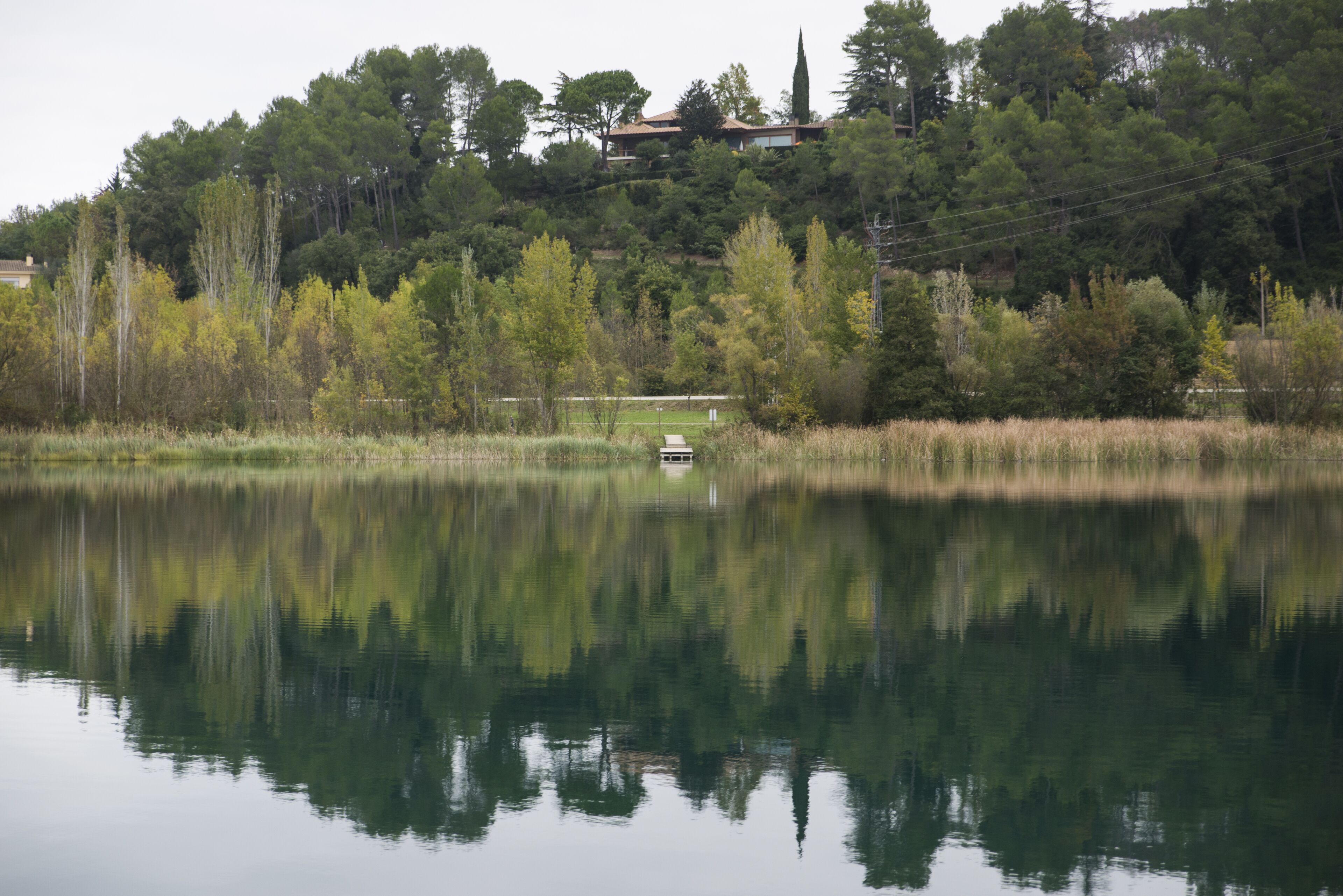 Un lago tranquilo con un bote pequeño, rodeado de árboles frondosos y una colina con una casa al fondo.