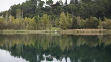 Un lago tranquilo con un bote pequeño, rodeado de árboles frondosos y una colina con una casa al fondo.