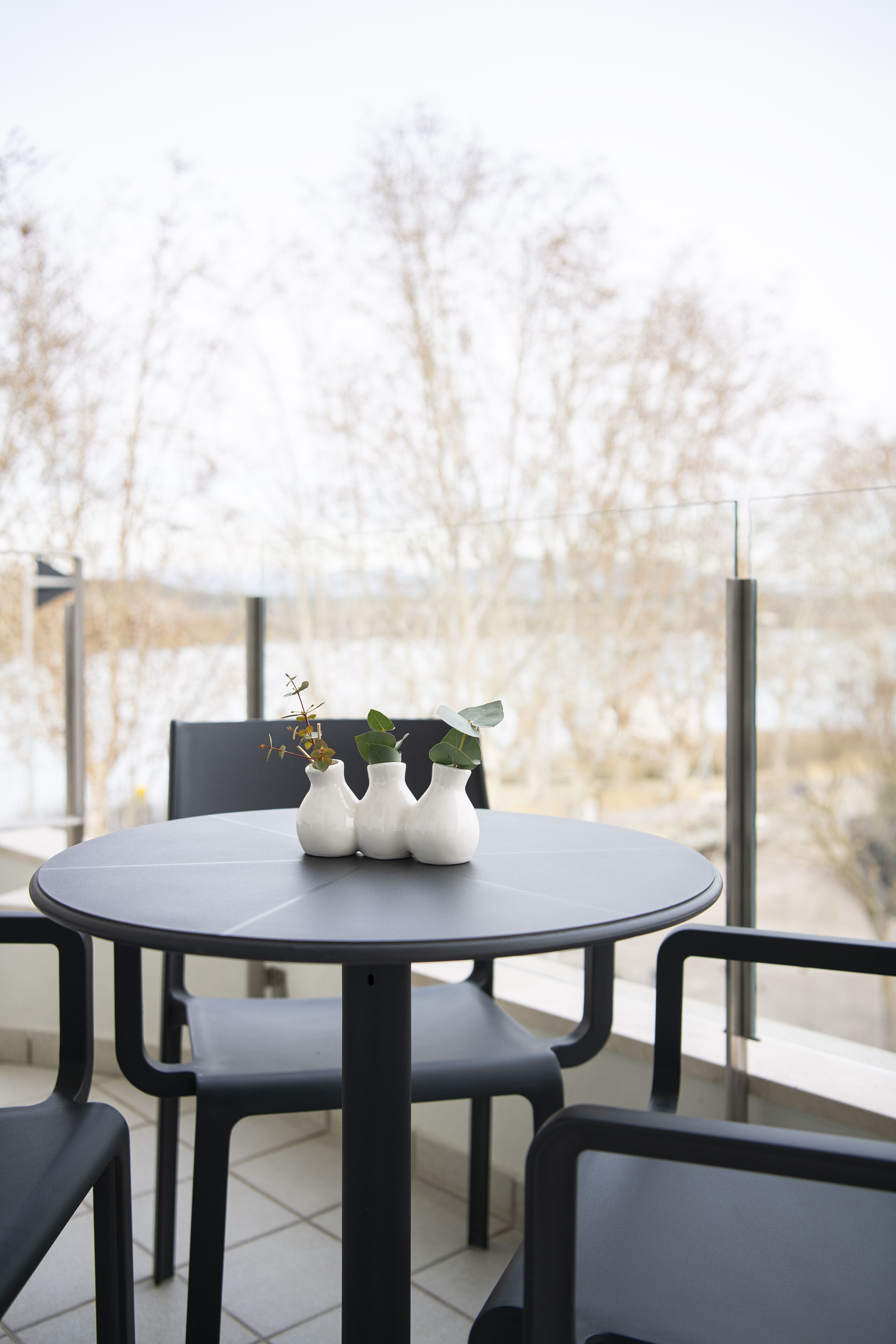 A round table with three white vases and a potted plant, surrounded by black chairs, on a balcony with a view of leafless trees.