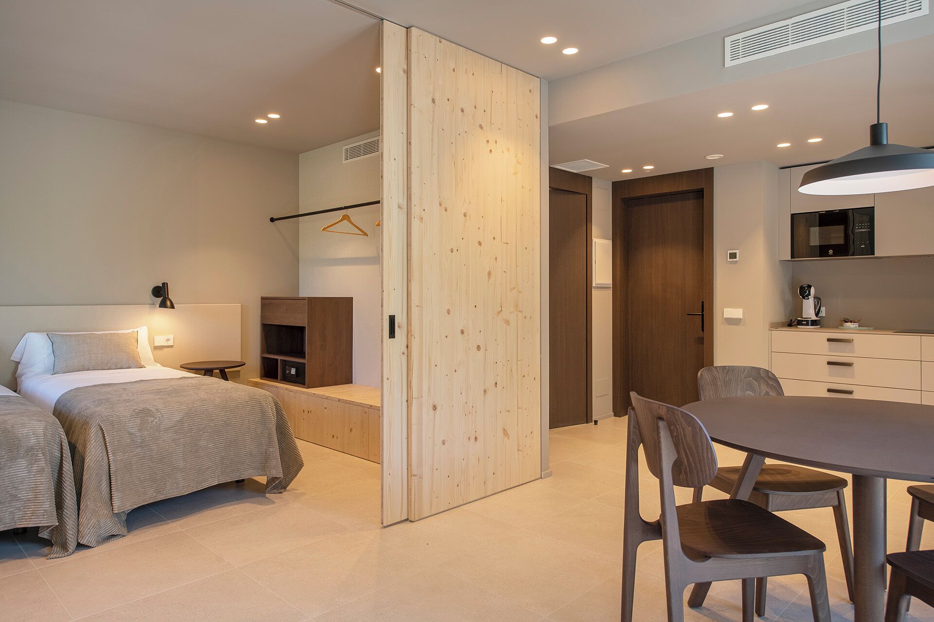 Camera d'albergo moderna con un letto, un comodino e un divisorio in legno. È presente un'area pranzo con tavolo e sedie.