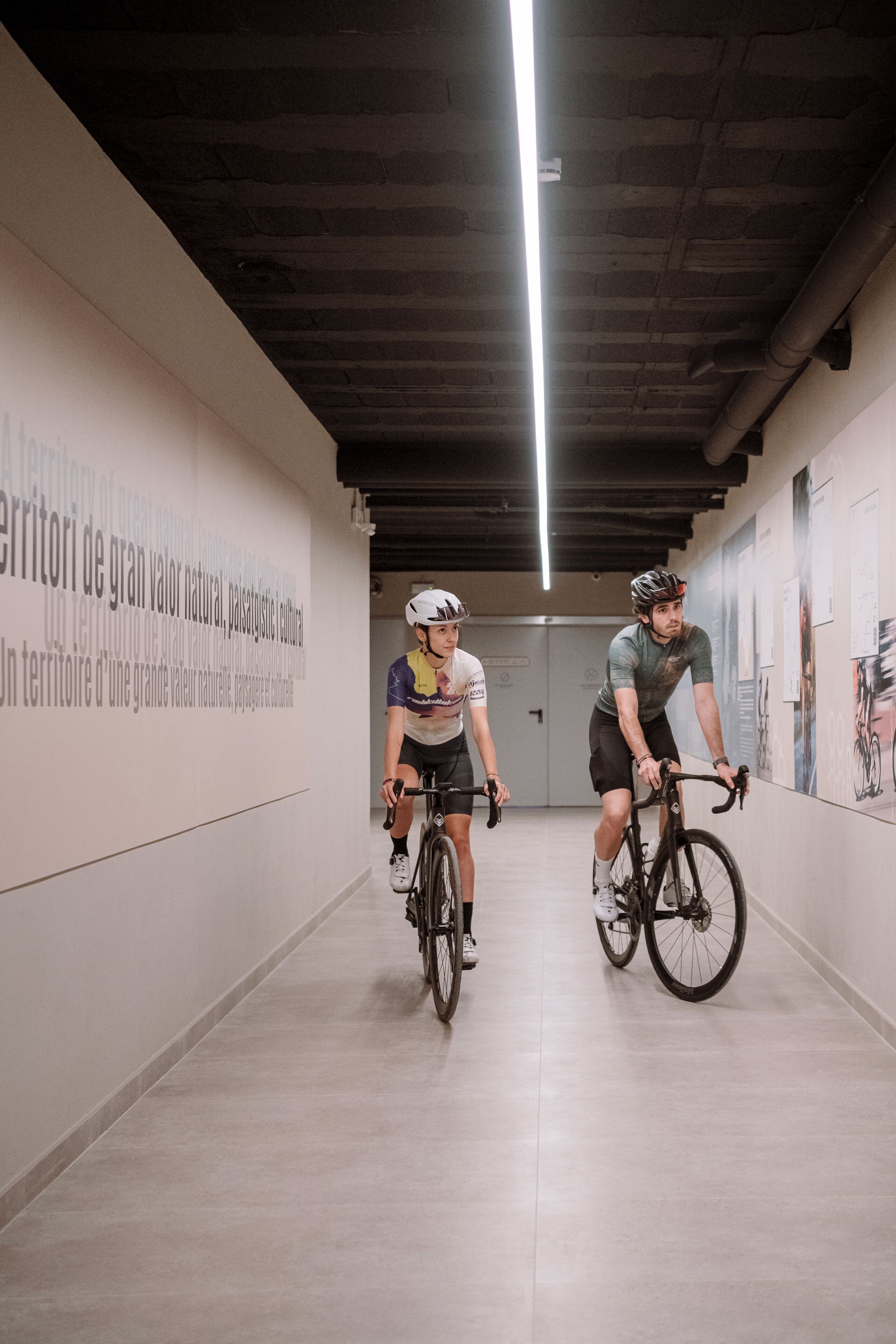 Two cyclists in a hallway with a tiled floor and a wall with text.