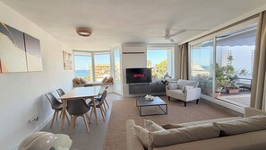 A modern living room with a flat-screen TV, a sofa, a dining table with chairs, and large windows offering a view of the sea.